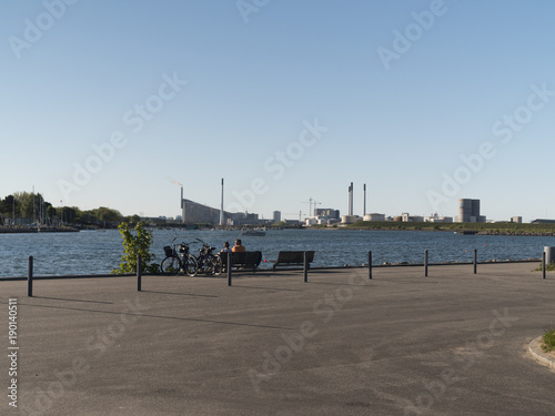 Pier with industrial buildings and people sittong on a bench enjoying the view ower the water and the blue sky