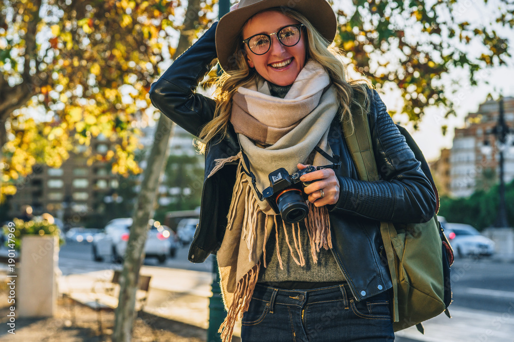 Sunny day, autumn.Young woman tourist, photographer, hipster girl dressed in hat and eyeglasses,sits on bench on city street and takes photo.Vacation, travel,adventure, sightseeing.Blurred background.