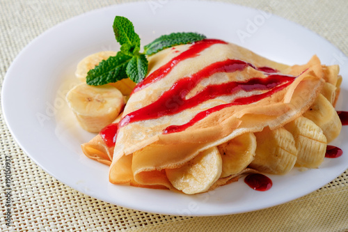 On a light table, the white plate with sliced bananas wrapped in pancakes. Pancakes are poured with jam and decorated with green mint leaves.