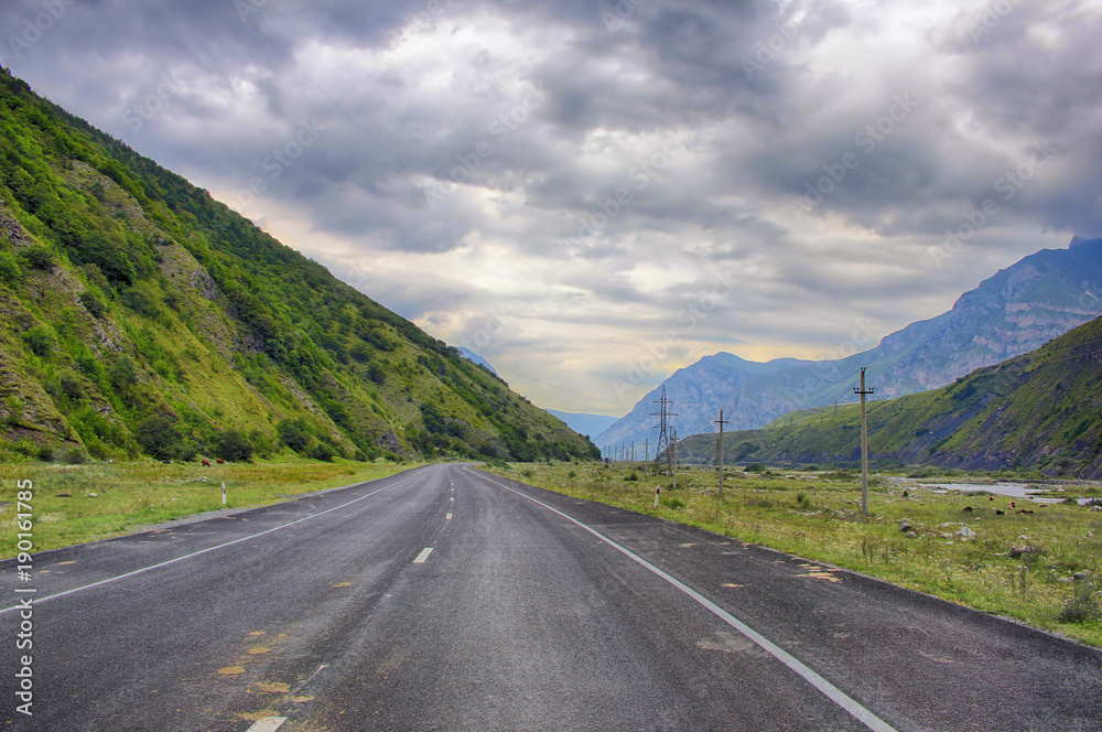 Highway running away through the mountain valley between high green ranges under cloudy sky.