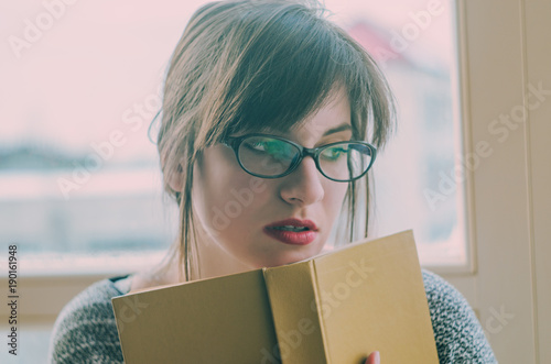 literature, education and people concept - young woman in glasses reading book at home