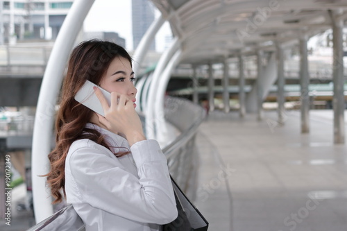 Attractive young Asian business woman talking on mobile smart phone and  holding document folder at outside office with copy space background. © tuaindeed