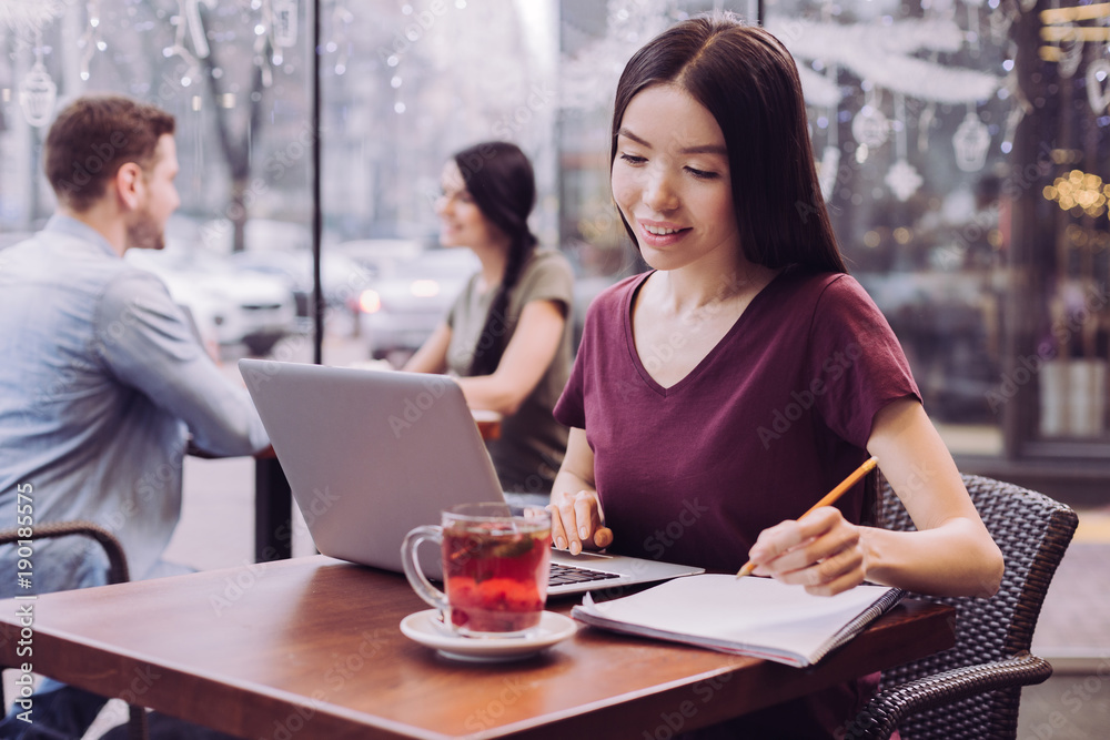 Useful information. Pensive appealing female student using laptop while looking at the screen and writing down information Useful information. Pensive appealing female student using laptop while looking at the screen and writing down information