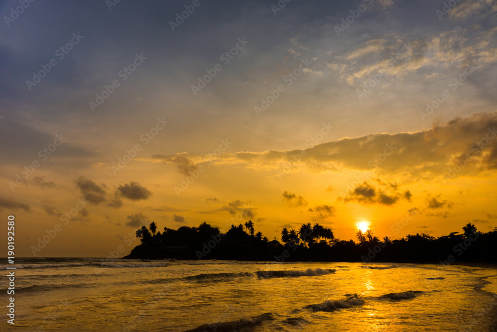 Indian ocean waves and dark silhouettes of palm trees on the horizon against beautiful colorful cloudy sky at sunset. Dewota beach, Sri Lanka.