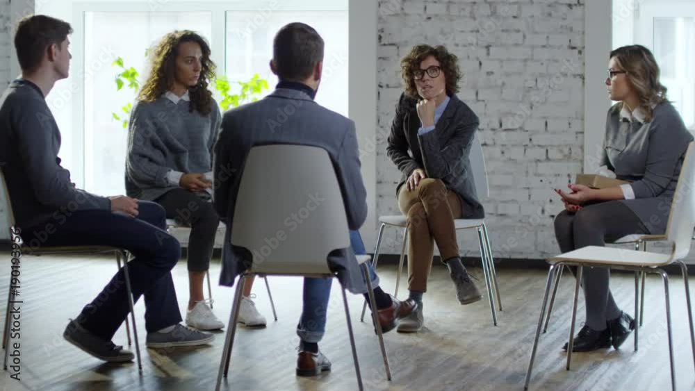 Wide shot of curly young woman sitting with crossed legs and discussing problems with male psychotherapist during support group meeting Wide shot of curly young woman sitting with crossed legs and discussing problems with male psychotherapist during support group meeting