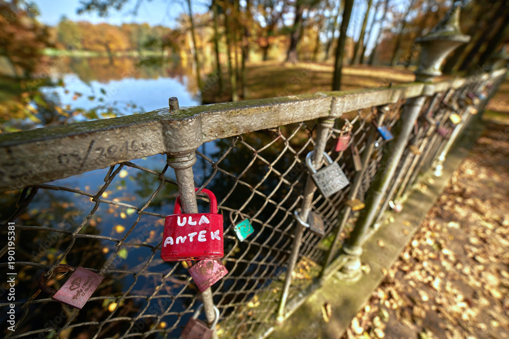 Słoneczny jesienny krajobraz - park w złotych kolorach, balustrada na moście miłości udekorowana kłódkami zakochanych par, w tle staw otoczony drzewami