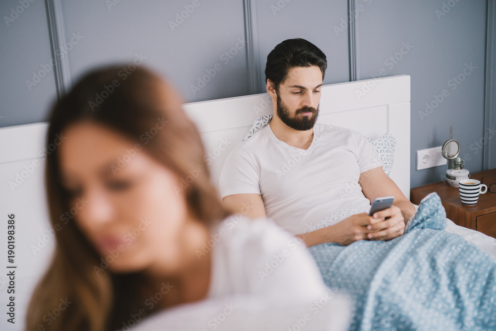 Bearded man looking at his phone while lying in bed with a young woman with brown hair.