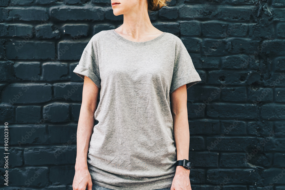 Summer day. Front view. Young millennial woman dressed in gray t-shirt is stands against dark brick wall. Mock up. Space for logo, text, image. Summer day. Front view. Young millennial woman dressed in gray t-shirt is stands against dark brick wall. Mock up. Space for logo, text, image.