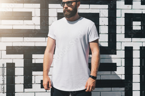 Summer day. Front view. Young bearded millennial man dressed in white t-shirt and sunglasses is stands against brick wall. Mock up. Space for logo, text, image. Instagram filter, film effect, bokeh