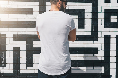 Summer day. Back view. Young bearded millennial man dressed in white t-shirt is stands against brick wall. Mock up. Space for logo, text, image. Instagram filter, film effect, bokeh effect.