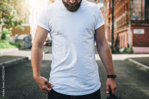 Summer day. Front view. Young bearded millennial man dressed in white t-shirt is stands on city street. Mock up. Space for logo, text, image. Instagram filter, film effect, bokeh effect.