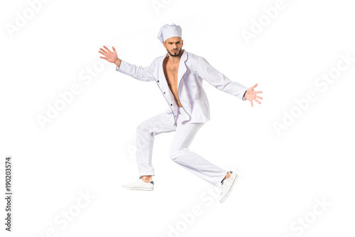 handsome young chef in uniform jumping and looking at camera isolated on white