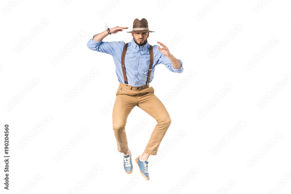 stylish young man in hat jumping and looking at camera isolated on white
