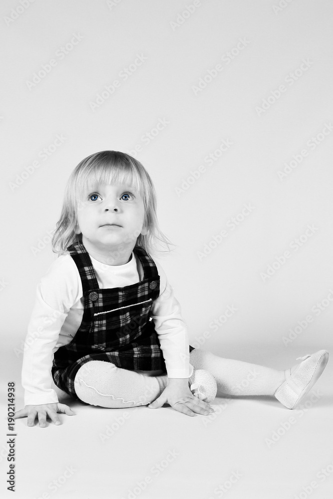 Naklejka premium Little blonde toddler girl with big Blue Eyes in red dress looking up.Sitting on the floor, white background. Portrait, close-up isolated , Black and White photo
