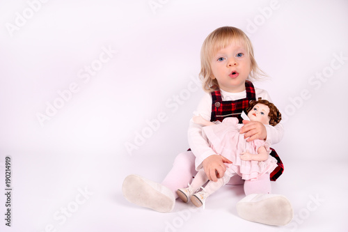 Little blonde toddler girl with big Blue Eyes in red  dress play with old doll. Sitting on the floor, white background. Portrait, close-up isolated 