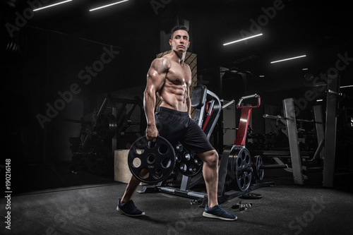 A young male bodybuilder does exercises with sports equipment, makes sit-ups with a barbell, in a modern gym. Weightlifting. On a dark background.