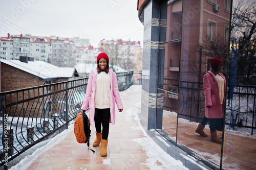 African american girl in red hat and pink coat with backpack at street of city against building on winter day. © AS Photo Family