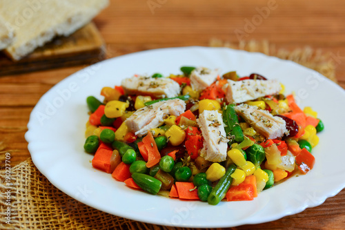 Chicken fillet and mixed vegetable stew on a white plate and a wooden table. Simple chicken vegetable stew. Stewed carrots  green beans  red beans  peas  corn  pepper and onions with chicken