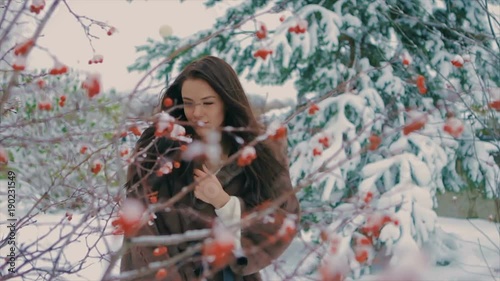 brunette stylish girl with bright makeup, near bush with red berries and Snow covered Christmas trees, in a luxurious brown fur coat walking in winter time slow motion