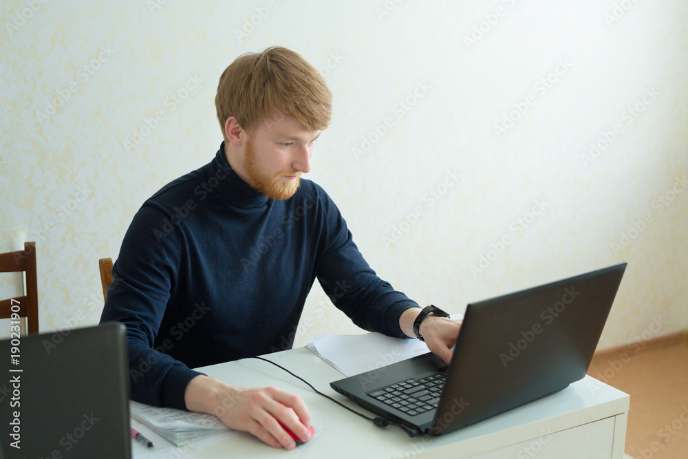 handsome young man with a red beard working in his office behind a laptop handsome young man with a red beard working in his office behind a laptop