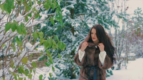 brunette stylish girl with bright makeup near green trees, in a luxurious brown fur coat walking in winter time slow motion
