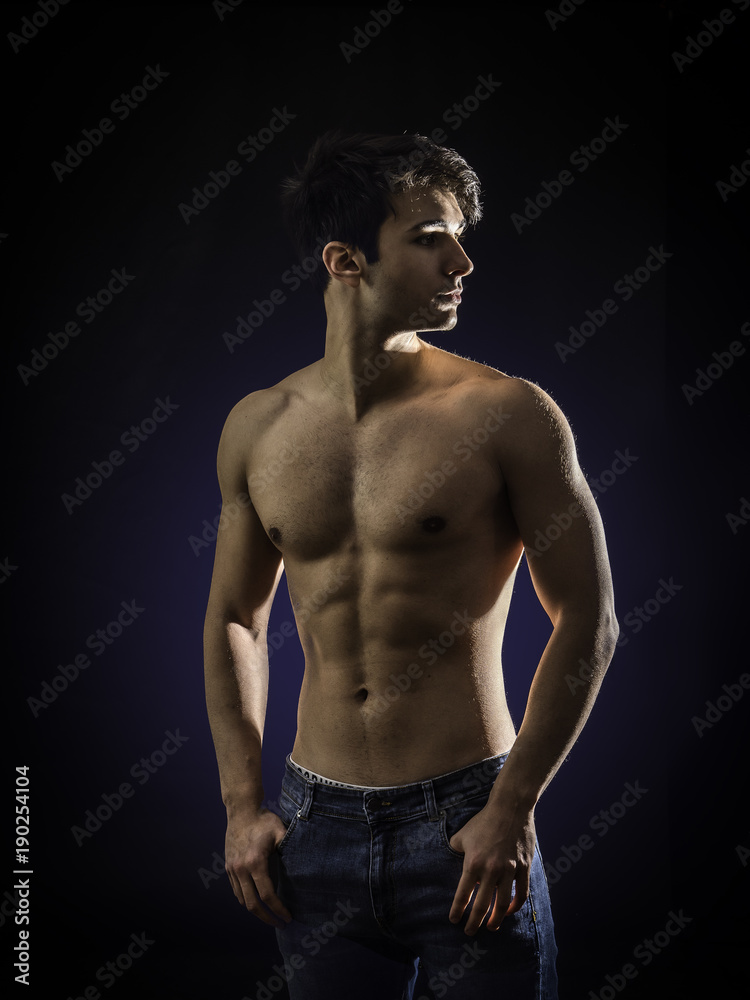 Handsome, fit muscular young man shirtless, wearing only jeans standing on black background, looking away to a side