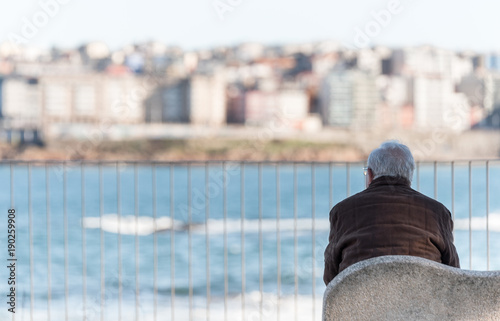 Elderly man sitting on a bench in the sun facing the sea