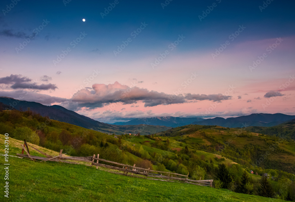 rural area at dusk with moon on cloudy sky. beautiful mountainous landscape with agricultural fields and wooden fences on grassy slopes in springtime