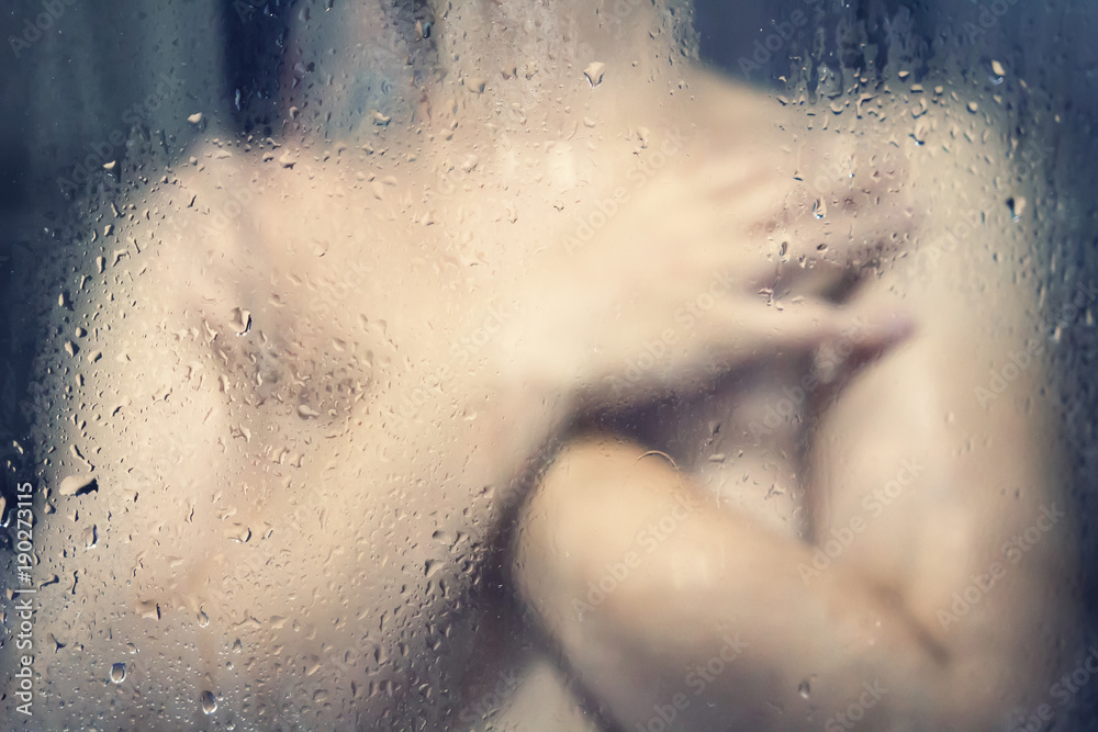 Naked girl in shower behind a wet glass with water droplets.