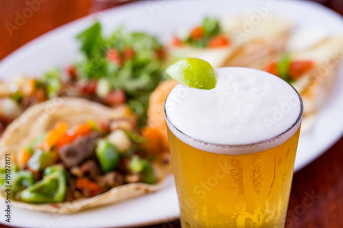 Cold beer with lime wedge and Mexican food dish in beef tacos in background.