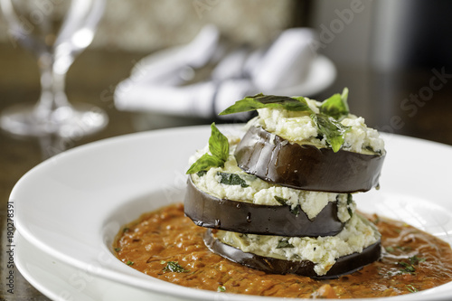 Stuffed eggplant in red marina sauce on white plate with glass and napkins in background.