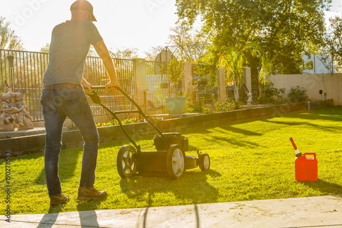 Man mowing  green lawn with push mower at mid-day with sun glowing in background.