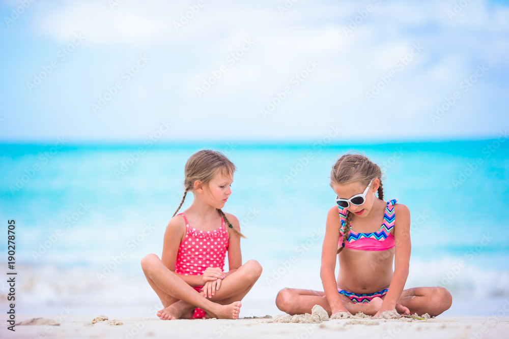 Adorable little girls playing with sand on the beach. Kids sitting in shallow water and making a sandcastle Adorable little girls playing with sand on the beach. Kids sitting in shallow water and making a sandcastle