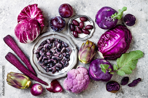 Raw purple vegetables over gray concrete background. Cabbage, radicchio salad, olives, kohlrabi, carrot, cauliflower, onions, artichoke, beans, potato, plums. Top view, flat lay.