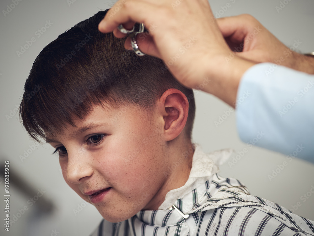 Fototapeta premium Barber is making haircut to cheerful boy in barbershop.