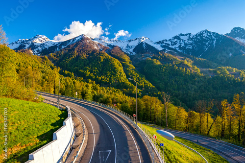 A winding modern asphalt road to the top of a resort in the bright yellow rays of the evening sun, dense forest and the Caucasian mountain range. Summer spring forest mountain landscape, Sochi Russia.