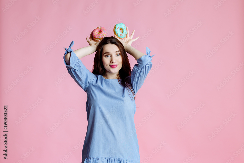 Portrait of young woman brunette with long beautiful hair in a blue dress with sweet donuts on pink background Portrait of young woman brunette with long beautiful hair in a blue dress with sweet donuts on pink background