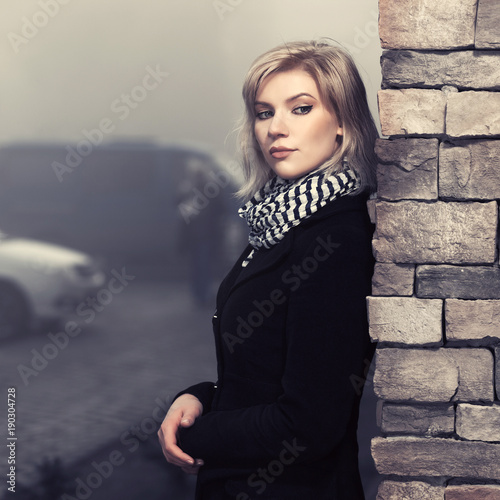 Fashion blond woman in black coat leaning on wall in city street