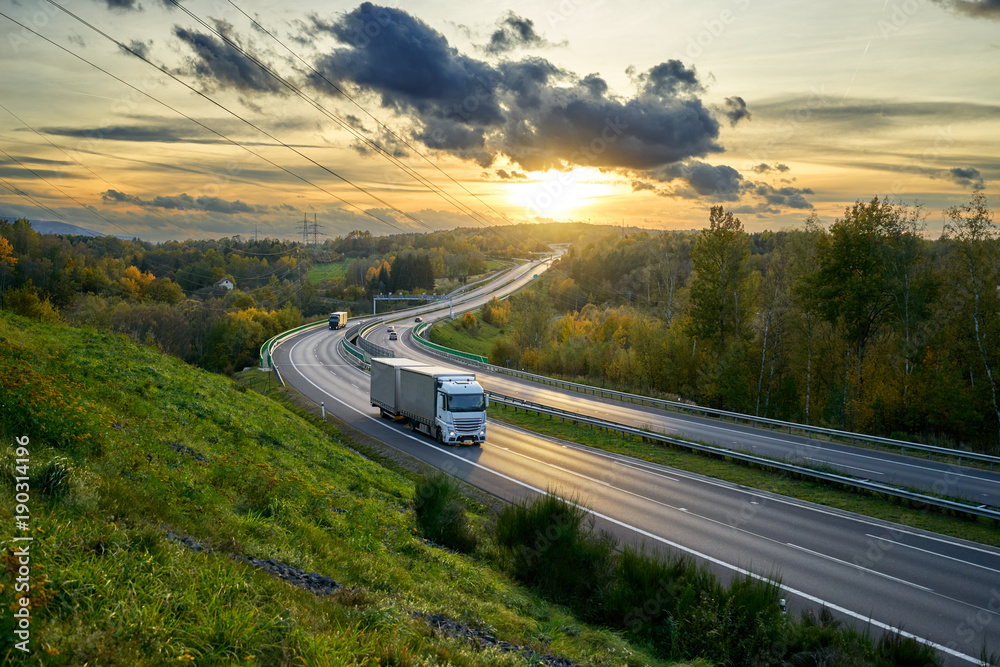 Trucks driving on the highway turning towards the horizon in an autumn landscape at sunset with dramatic clouds. Electronic toll gate in the middle.