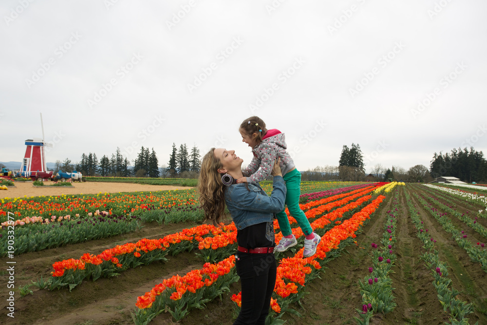 Portrait Close-up of Four Years Old Cute Girl Is Hugging Her Beautiful Young Mother, Looks at Camera and Smiling, Tulip Field at the Background, early spring, Tulip Farm, Oregon USA