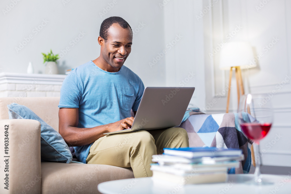 Modern entertainment. Happy joyful nice man smiling and using laptop while sitting on the sofa Modern entertainment. Happy joyful nice man smiling and using laptop while sitting on the sofa