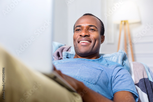 Rest at home. Joyful cheerful nice man smiling and looking at the laptop screen while resting at home