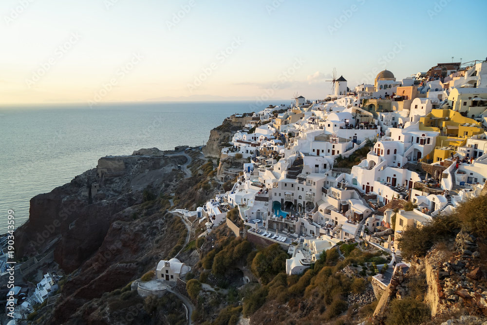 Panoramic scene in sunset light  of Oia windmill and white building townscape along island natural mountain facing vast ocean with soft cloud and light blue sky background