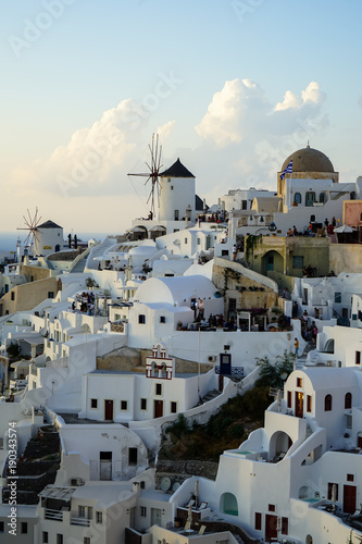 Evening light scene of Oia windmill and white building townscape along island mountain facing ocean with soft cloud and light blue sky background
