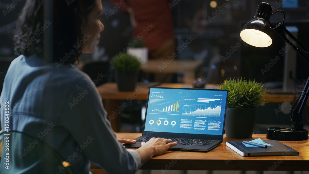 Female Analyst at Her Desk Works on a Laptop Showing Statistics, Graphs and Charts. She Works on the Wooden Table in Creative Office. Over the Shoulder Footage. Female Analyst at Her Desk Works on a Laptop Showing Statistics, Graphs and Charts. She Works on the Wooden Table in Creative Office. Over the Shoulder Footage.