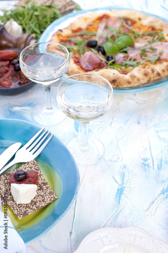 Two glasses of white wine. Pizza with prosciutto and olives. Green salad. Servian lunch, on a light background with punctuated spots. Free space for text or a postcard.