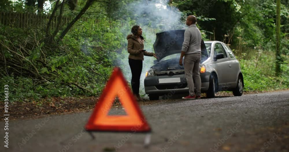 4K Stranded couple arguing in front of broken down car on quiet country road. Red warning triangle in foreground. Slow motion.