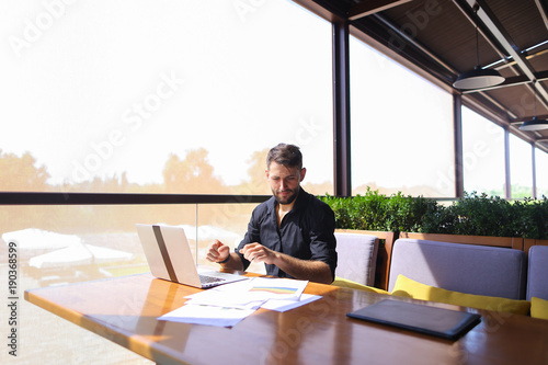 Tired quantity surveyor working at cafe table with diagram and statistic documents. Persistent man dressed in classic black shirt sitting at sofa near green indoor plants. Concept of  preparin photo