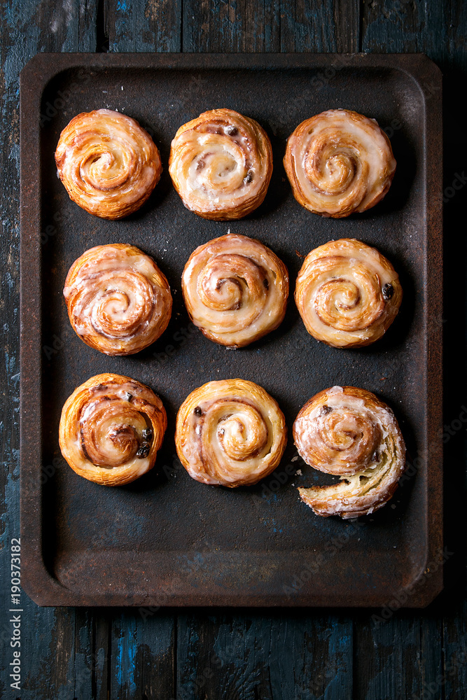 Homemade glazed puff pastry cinnamon rolls with custard and raisins on oven tray over old dark blue wooden background. Top view. Rustic style