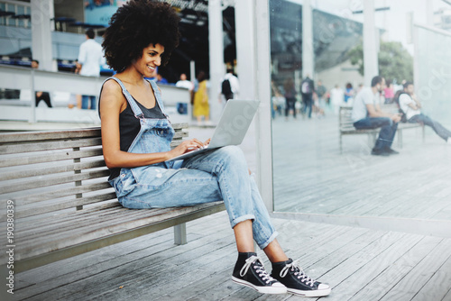 young black female IT specialist working remotely on a new project using a public network wifi while sitting on a wooden bench on the city street. Pretty mixed race girl using laptop computer for work